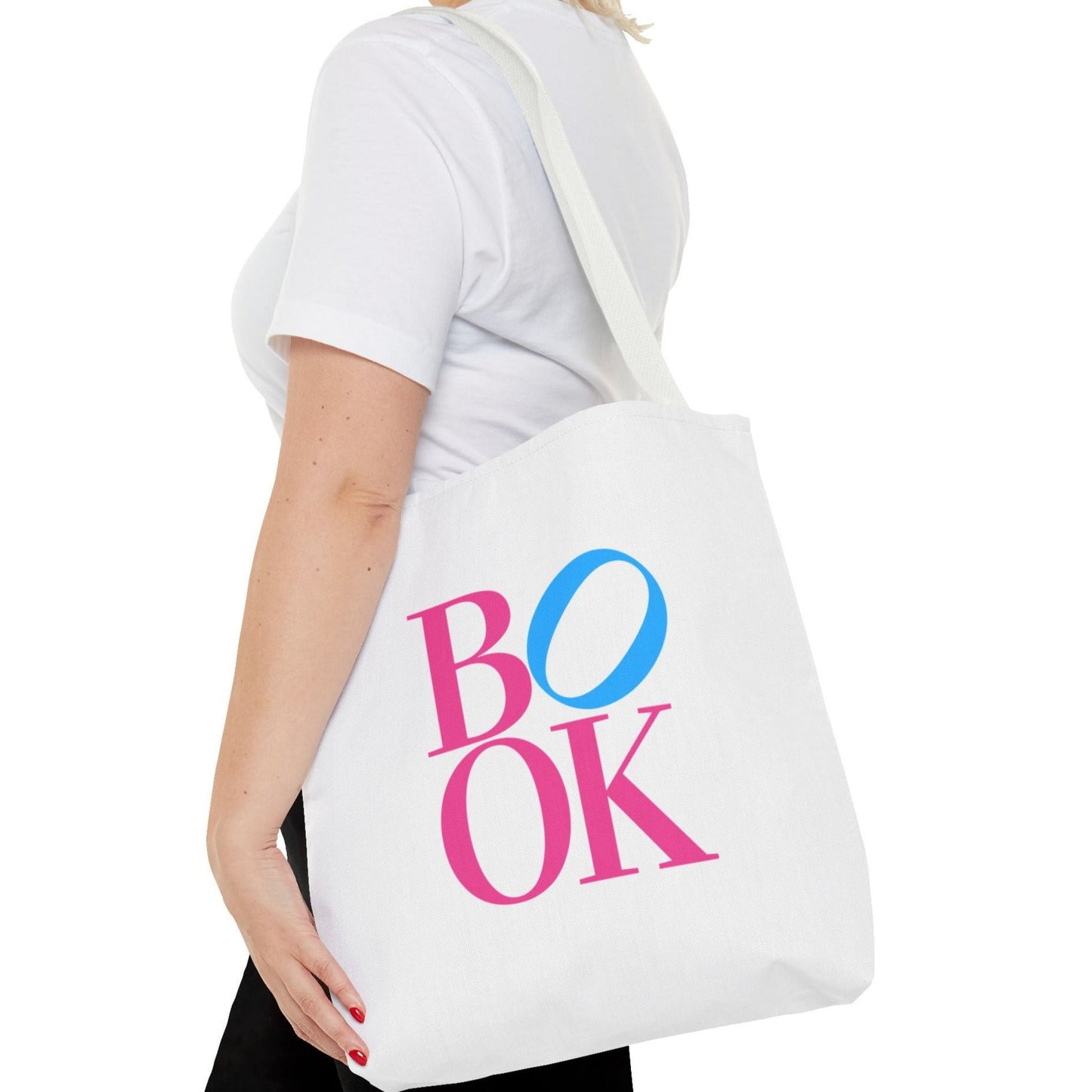 White tote bag with 'BOOK' printed in pink and blue held by a person wearing a white shirt.