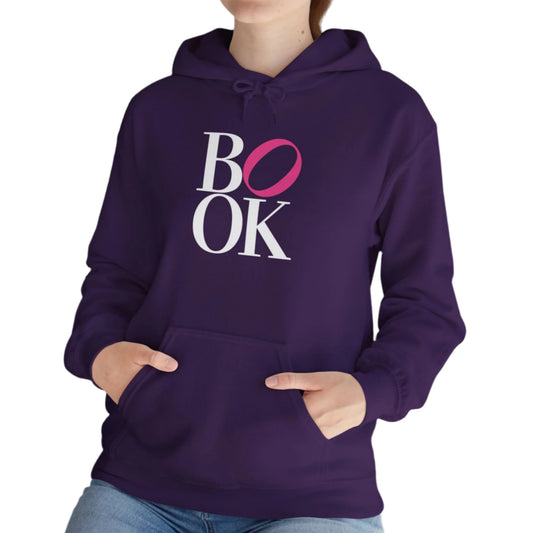 Person wearing a purple hoodie with 'BOOK' printed on it, standing indoors next to a plant.